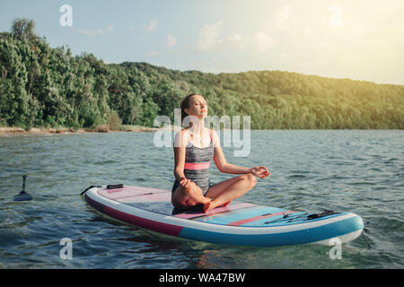 Medioevo donna caucasica la pratica dello yoga sulla paletta sup tavola da surf al tramonto. Femmina facendo stretching allenamento sul lago di acqua. Moderno tanga individuali Foto Stock