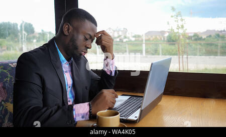 Afro American imprenditore tenta di digitare sul computer in seduta cafe. Uomo nero funziona su laptop ma si è rotto e lui è nervoso e battito sui tasti. Egli ha un computer problemi. La carica della batteria è bassa. Foto Stock