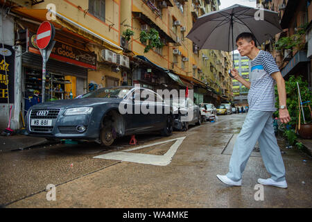 17 agosto 2019, Cina, Hong Kong: un uomo cammina con un ombrello attraverso una strada nel distretto di Kowloon, dove un auto senza ruote anteriori sorge in corrispondenza di un suo lato. Foto: Gregor Fischer/dpa Foto Stock