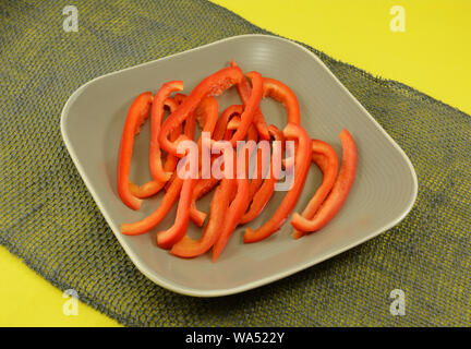 Sottili fette di crudo fresco peperone rosso sulla piastra sulla tabella di tessuto runner sulla tovaglia giallo Foto Stock