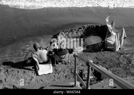 Un giovane seduto sulla spiaggia rilassarsi leggendo un libro e guardando il mare dal litorale sabbioso Foto Stock