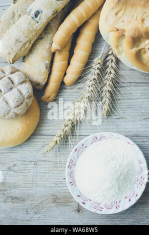 Diversi tipi di Italiano tradizionale di pane appena sfornato, vari tipi di pane e panini con ciotola di farina a fontana sul tavolo di legno dello sfondo. Rustico stile vintage. Vista dall'alto. Foto Stock