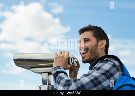 Vista laterale porta caratteristica di entusiasti turista maschio utilizzando un binocolo fisso alla piattaforma di osservazione durante le vacanze, spazio di copia Foto Stock