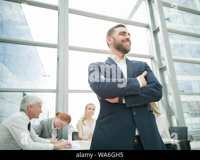 Fiducioso giovane imprenditore in piedi con le braccia incrociate sorridendo , squadra di suoi colleghi seduti a tavola in background Foto Stock