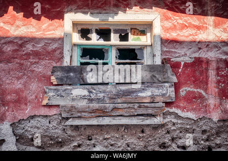 Finestra con vetro rotto e bloccate chiuse con assi sulla parete rossa di un abbandonato shanty house Foto Stock