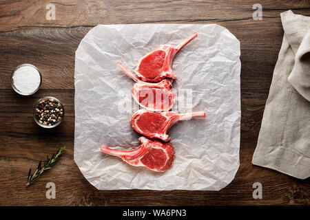 Fette di materie agnello costolette di nervatura sul bianco di carta di cottura e tavolo in legno. Decorate con sale e pepe, carne forcella e igienico. Vista aerea. Foto Stock