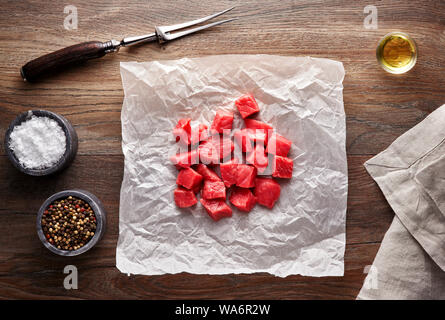 Un trito di materie bovine cubetti di carne per spezzatino di carne in bianco di carta di cottura e tavolo in legno. Decorate con sale e pepe, carne forcella e igienico. Vista aerea. Foto Stock