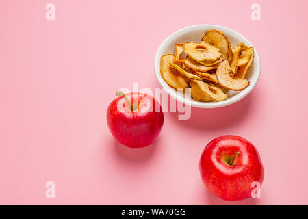 Secche mele chips in ceramica bianca ciotola con fresche Mele rosse su sfondo rosa Foto Stock
