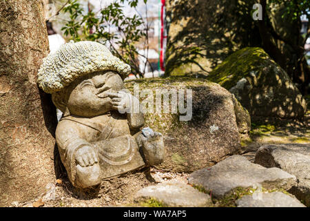 Giappone, Miyajima. Daisho-nel tempio. Rakan piccola statua di un monaco buddista, con rosa cappello di lana, il dito contro il labbro, sembra che il pensiero è duro. Foto Stock