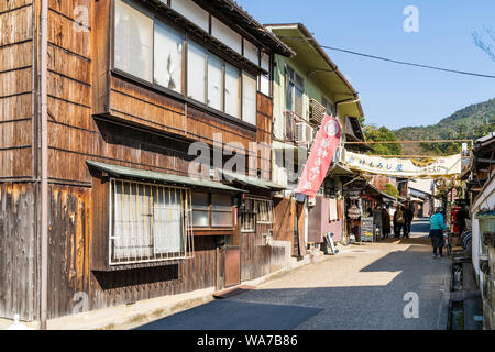 Il Giappone, l'isola di Miyajima. Piccola strada laterale con negozi e case che conduce su per la collina con alcune persone a piedi. Non occupato. Sullo sfondo le montagne della foresta. Foto Stock