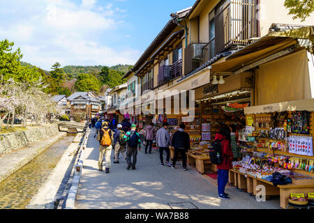 Il Giappone, l'isola di Miyajima. I turisti a piedi oltre la riga di acquisto di souvenir negozi lungo la strada con terrapieno e piccolo ruscello sull'altro lato. Fiori di Ciliegio. Foto Stock