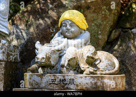 Giappone, Miyajima. Daisho-nel tempio. Piccola pietra Jizo statua della seduta monaco buddista con cappello giallo, lato seduto sulla sella sitting bull, Buffalo. Foto Stock