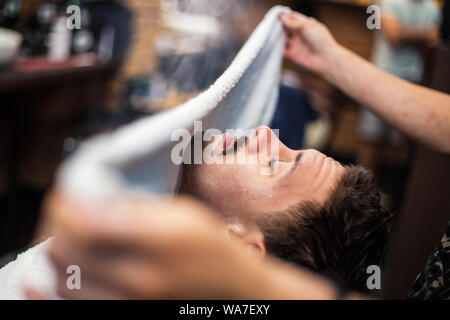 Barbiere copre il volto di un uomo con una salvietta calda. Rito della rasatura della barba con impacchi caldi e freddi in un vecchio stile di barbiere. Foto Stock