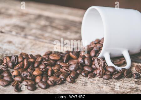 Primo piano di una tazza di ceramica bianca con chicchi di caffè versando fuori di esso su una superficie di legno Foto Stock