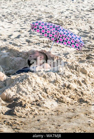Un uomo stanco si rilassa in un fresco foro scavato nella sabbia e sotto l'ombra di una polka dot ombrello Foto Stock