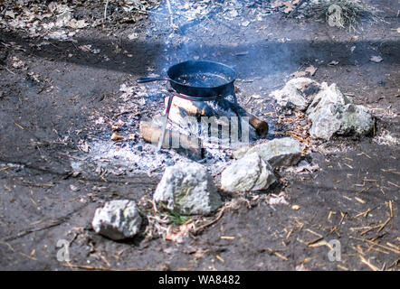 Una padella in ghisa si siede su un supporto su un ardente fuoco di campo nei boschi Foto Stock