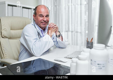 Ritratto sul sorridente medico esperto in labcoat seduto al tavolo per ufficio Foto Stock
