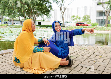 Sorridente giovane donna asiatica in abito islamico puntando verso qualcosa di divertente per mostrare al suo amico ridere seduta vicino da Foto Stock