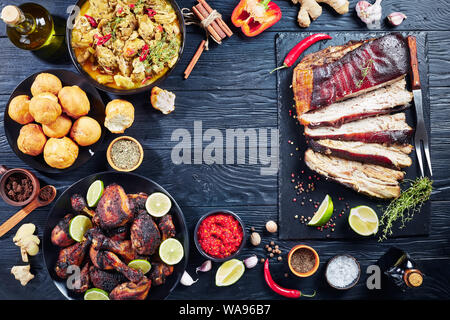 Set di piatti caraibici, jerk pancetta di maiale, pollo al curry, gnocco fritto, arrosto di pollo Cosce e fusi su piastre su tavoli in legno nero, vie Foto Stock