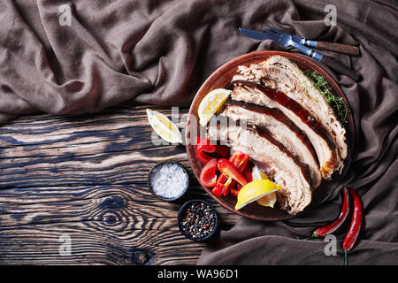 Fette di carne di maiale a scatti ventre con spicchi di limone e peperone su una piastra di argilla su un tavolo in legno con panno marrone, close-up Foto Stock