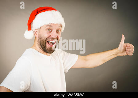 Uomo Barbuto che indossa santa claus hat dando pollice in alto segno ok, sul grigio con copia spazio. Tempo Christmes. Foto Stock