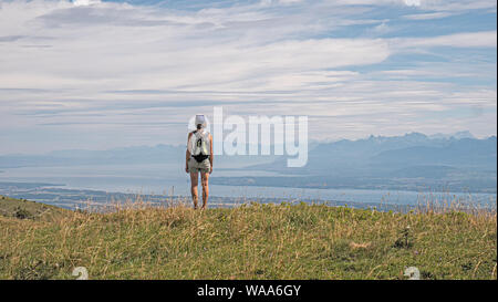 Una femmina solitario escursionista sorge in cima a una montagna guardando alla vista di un grande lago alpino circostante e delle vette alpine. Foto Stock