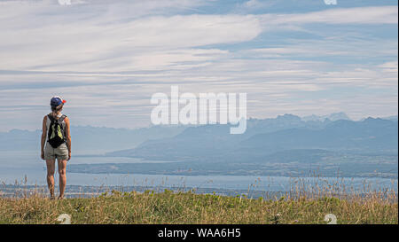 Una femmina solitario escursionista sorge in cima a una montagna guardando alla vista di un grande lago alpino circostante e delle vette alpine. Foto Stock