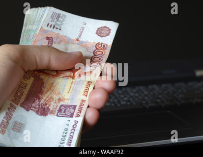 Prestiti online. Man mano che tiene un fascio di russo di denaro. Laptop in background. Banconote 5000 rubli. Compensazione freelance in Russia. Foto Stock