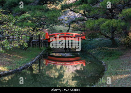 Un ponte ad arco rosso luminoso e riflesso così come i fiori di ciliegio sullo sfondo al Giardino di Ritsurin a Shikoku, Giappone Foto Stock