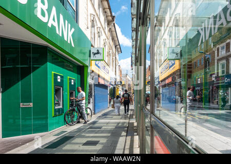 Un uomo utilizza una macchina ATM a Slough High Street ramo di Lloyds Bank. La scena si vede riflessa in un vetro fermata. Foto Stock