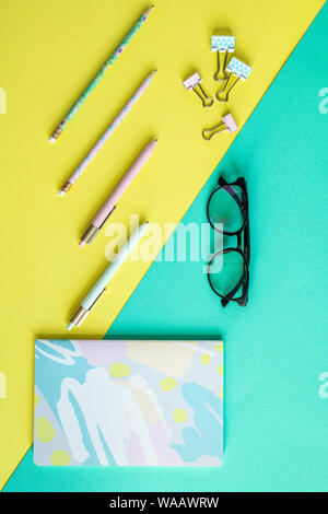 Panoramica delle penne, clip, notebook e occhiali su giallo e blu Foto Stock