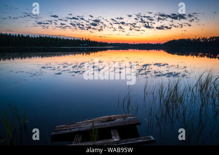 Tramonto colorato e la solitudine a distanza lago finlandese con bellissimi riflessi di nuvole e scura foresta boreale e molo vecchio in primo piano, finlan Foto Stock