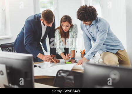 Team di tre giovani architetti che lavorano e comunicare durante l'analisi di schemi di un nuovo progetto in ufficio. Foto Stock