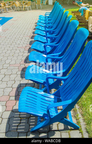 Estate recreation area dalla piscina all'aperto, moderno vuoto blu in plastica Sedie a sdraio stand in una fila sulla pavimentazione in lastre su un estate calda giornata di sole, verti Foto Stock