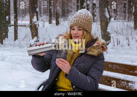 Ragazza seduta su una panchina nel parco invernale e versa se stessa il tè caldo da un thermos Foto Stock