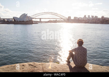 Uomo con bun, sitted sul bordo dell'acqua, fissando il Porto di Sydney Foto Stock