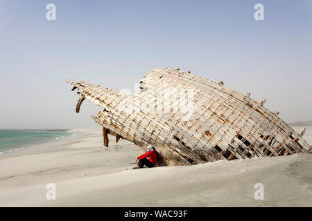 L uomo indossa foulard di testa seduto sotto 'la barca persa', relitto di dhow su una spiaggia di sabbia bianca sulla costa est isola Masirah, Oman Foto Stock