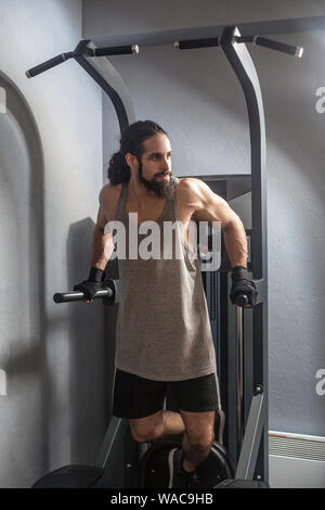 A piena lunghezza Ritratto di fiducioso giovane adulto uomo forte con lunghi capelli ricci che lavora fuori in palestra, appeso sulla barra orizzontale con le mani, facendo tirare ups Foto Stock