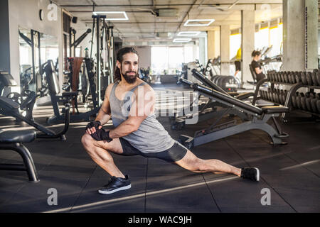 Vista laterale del bel giovane adulto sportman atleta con lunghi capelli ricci che lavora fuori in palestra, squating su un ginocchio , stretching dopo la formazione, facendo ex Foto Stock