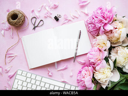 Piatto di blogger laici o area di lavoro freelance con una tastiera, notebook, peonie rosa su una rosa di sfondo in calcestruzzo Foto Stock