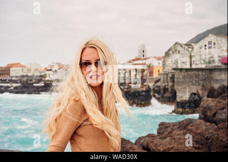 Attraente donna bionda in occhiali da sole contro la città sulla costa rocciosa con blu oceano onde sullo sfondo e lo sguardo lontano, Tenerife, Spagna Foto Stock