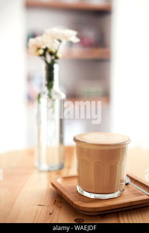 Cappuccino con schiuma spumeggiante, caffè nella tazza di vetro è sul tavolo con il vaso con fiori di colore bianco. Foto Stock