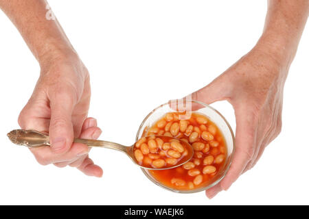Un senior donna anziana prende un cucchiaio di fave con salsa di pomodoro da una ciotola. Isolato su bianco studio closeup Foto Stock