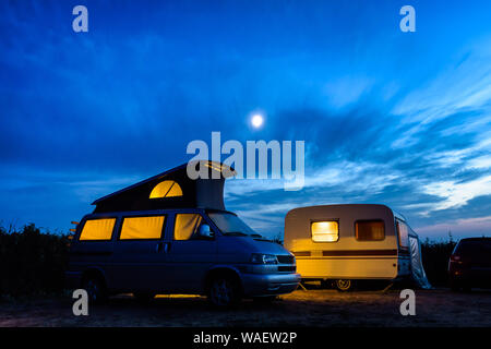 Un piccolo camper parcheggiato a fianco di un'annata roulotte in un campeggio, sia illuminato dall'interno al calar della sera con la luna risplende in un cielo tempestoso. Foto Stock
