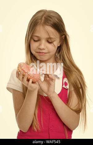 Ragazza con lunghi capelli biondi holding yummy deserto. Kid picking negli zuccherini colorati off ciambella. Sweet Tooth in abito rosa isolato su sfondo bianco. Ragazza mangiare ciliegina da bagel, zucchero coma concetto. Foto Stock