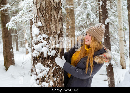 Allegro ragazza in un maglione caldo e berretto lavorato a maglia in un parco invernale la riproduzione di nascondersi dietro a un tronco di albero Foto Stock
