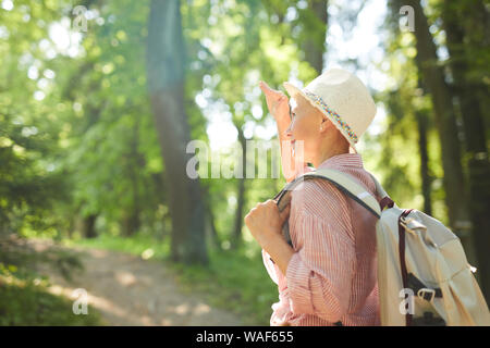 Donna matura in hat con zaino dietro la schiena che guarda lontano mentre si cammina nel bosco in estate Foto Stock