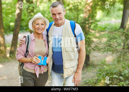 Ritratto di coppia senior di viaggiatori con zaino e binocolo sorridente in telecamera mentre in piedi insieme al parco all'aperto Foto Stock