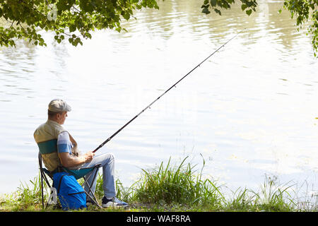 Uomo maturo di relax all'aperto al lungolago egli seduti su una sedia con la canna da pesca e la pesca Foto Stock