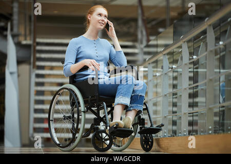 I Capelli rossi ragazza seduta in sedia a rotelle e parlando al telefono cellulare al moderno centro commerciale Foto Stock